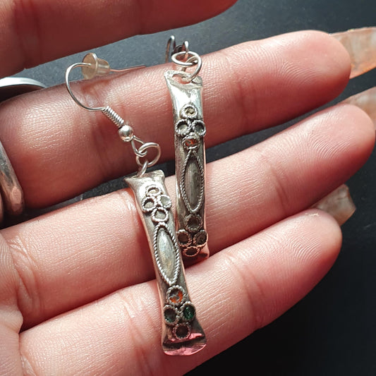 A close-up image of a pair of silver-tone dangle earrings with a curved design. The earrings feature intricate floral detailing, including small circular flowers and a marquise-shaped center with rope-like edging.